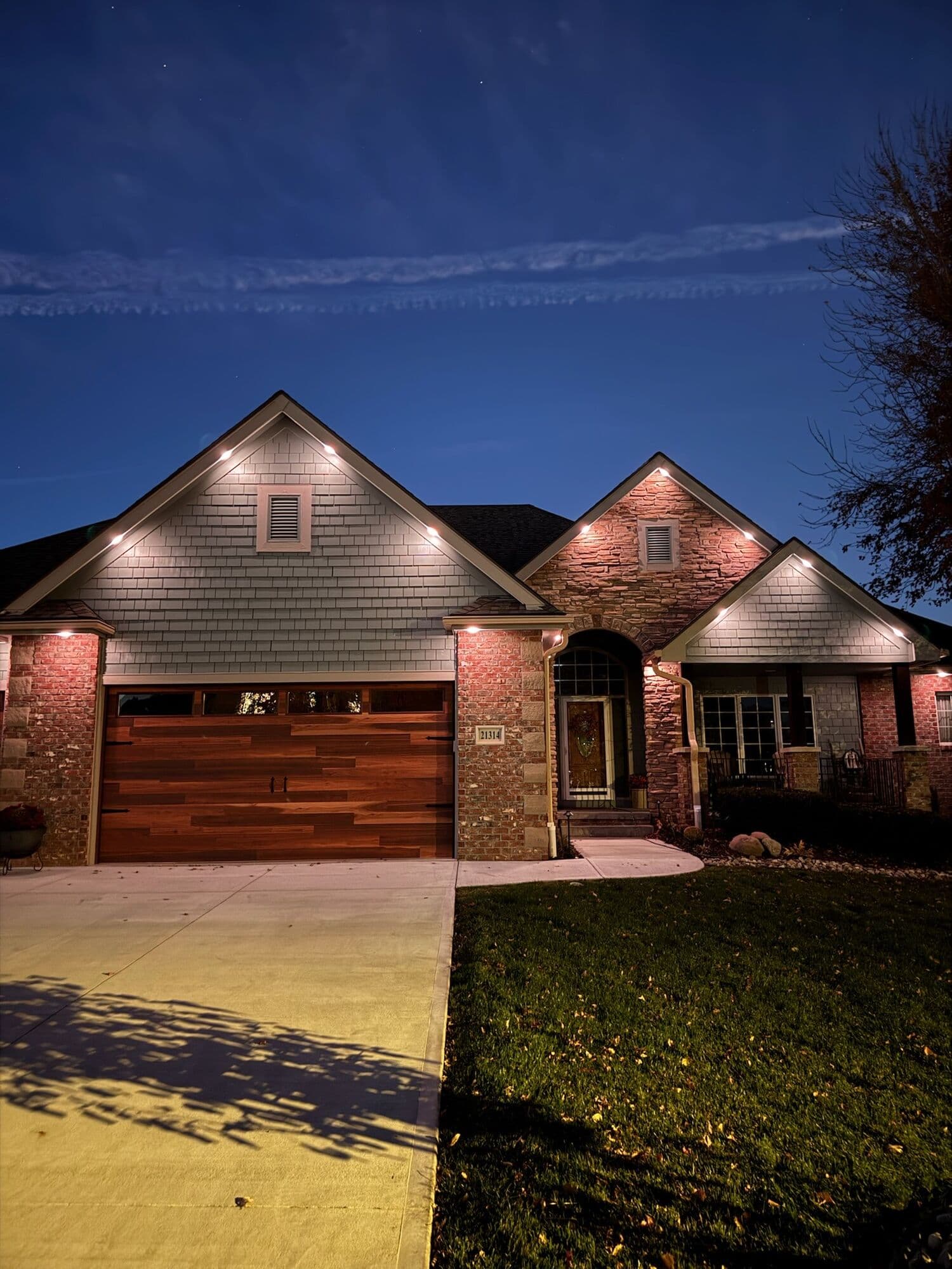 Warm white downlights under the eaves and gable peaks of a shake and brick Omaha home at blue hour, the spacing between points readable along each run